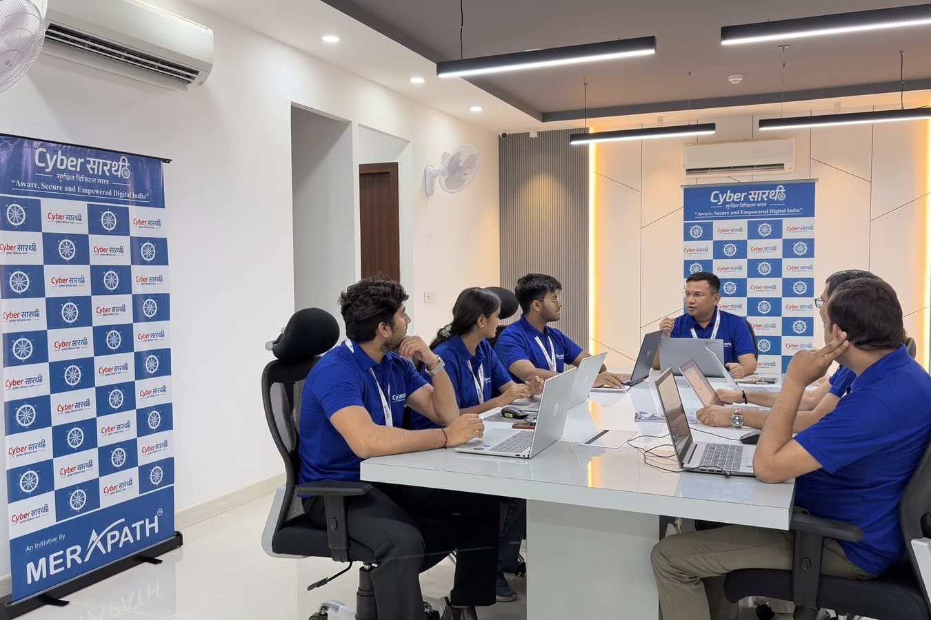 The Cyber सारथी team working at a table between two Cyber सारथी / MeraPath step-and-repeat banners.
